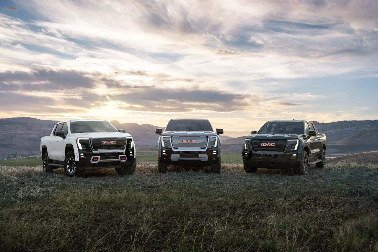 Three GMC Sierra EV pickups parked side by side on a grassy ridge at sunset