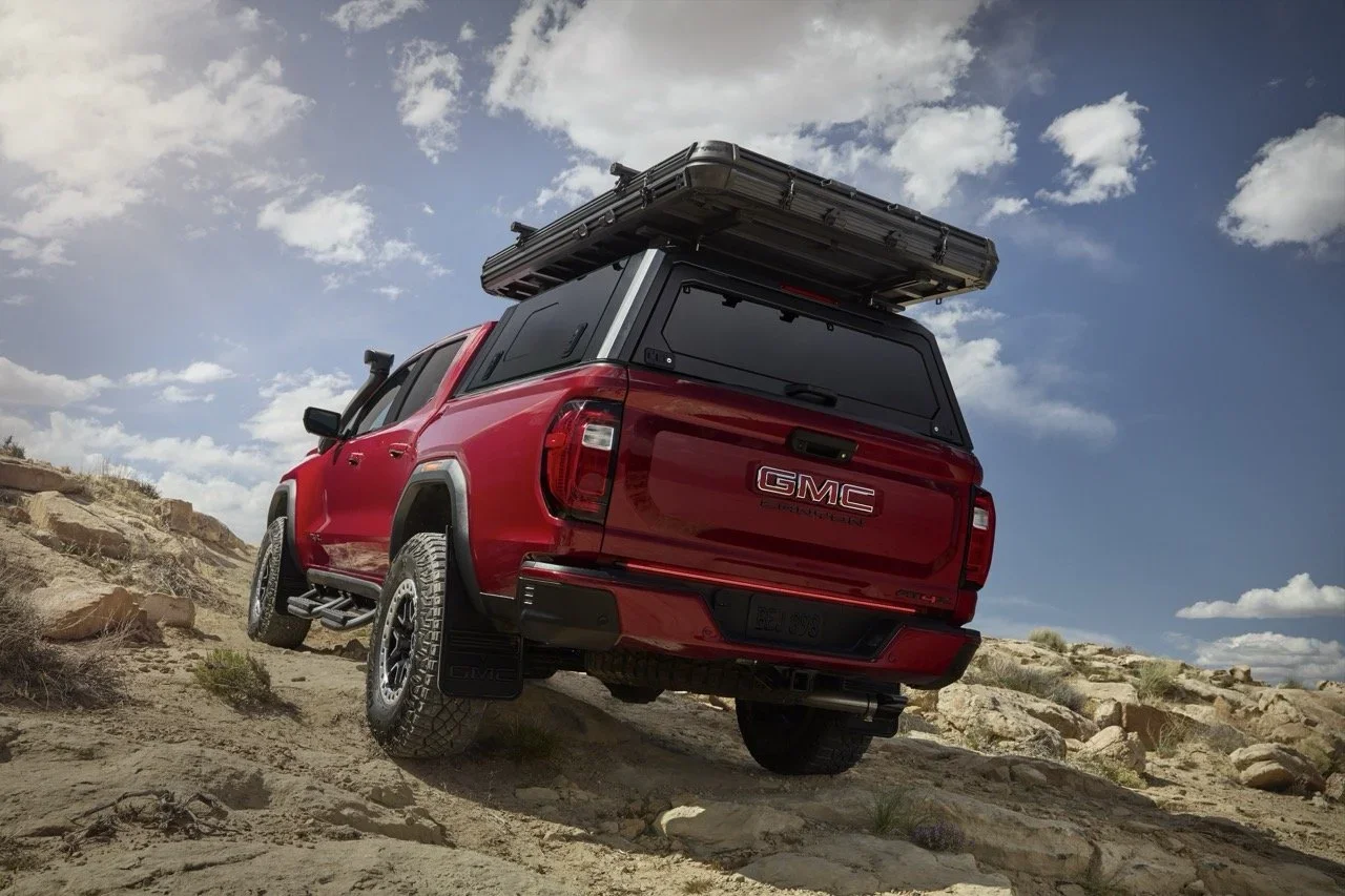 Red GMC Canyon climbing rocky terrain, rear view with bed cap and roof rack
