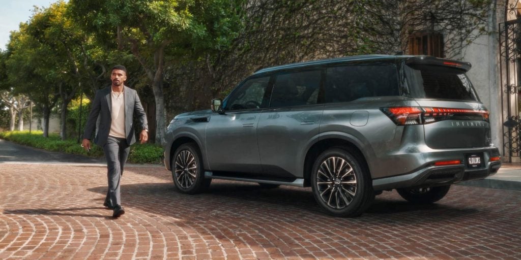 A well-dressed man walks along a tree-lined cobblestone driveway beside a sleek gray luxury SUV parked near a vine-covered building.