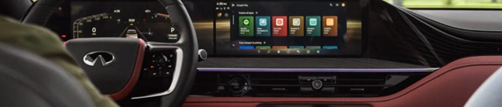 Driver’s view of an Infiniti car interior with steering wheel, digital dashboard, and wide infotainment touchscreen displaying apps.