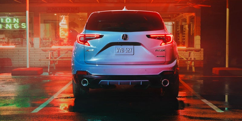 A white Acura RDX SUV is parked in a wet, neon-lit parking lot at night, viewed from the rear, with a restaurant in the background.