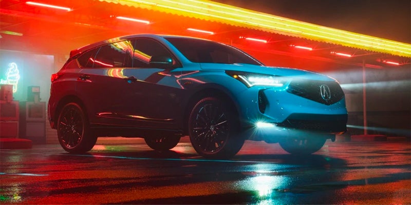 A blue 2025 Acura RDX SUV parked on a wet street with colorful neon lights reflecting off the surface.