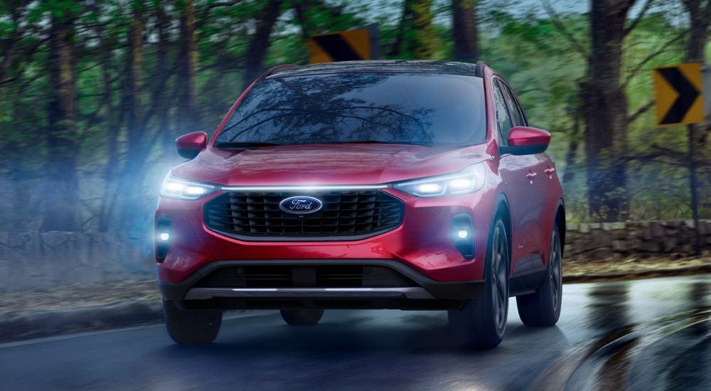 Red 2026 Ford Escape driving on a paved road with headlights on.