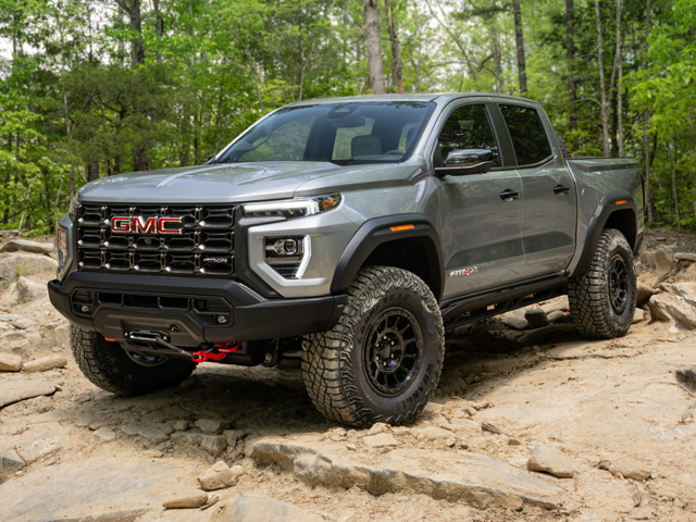 A grey 2024 GMC Canyon parked on rugged terrain with a tree line in the background