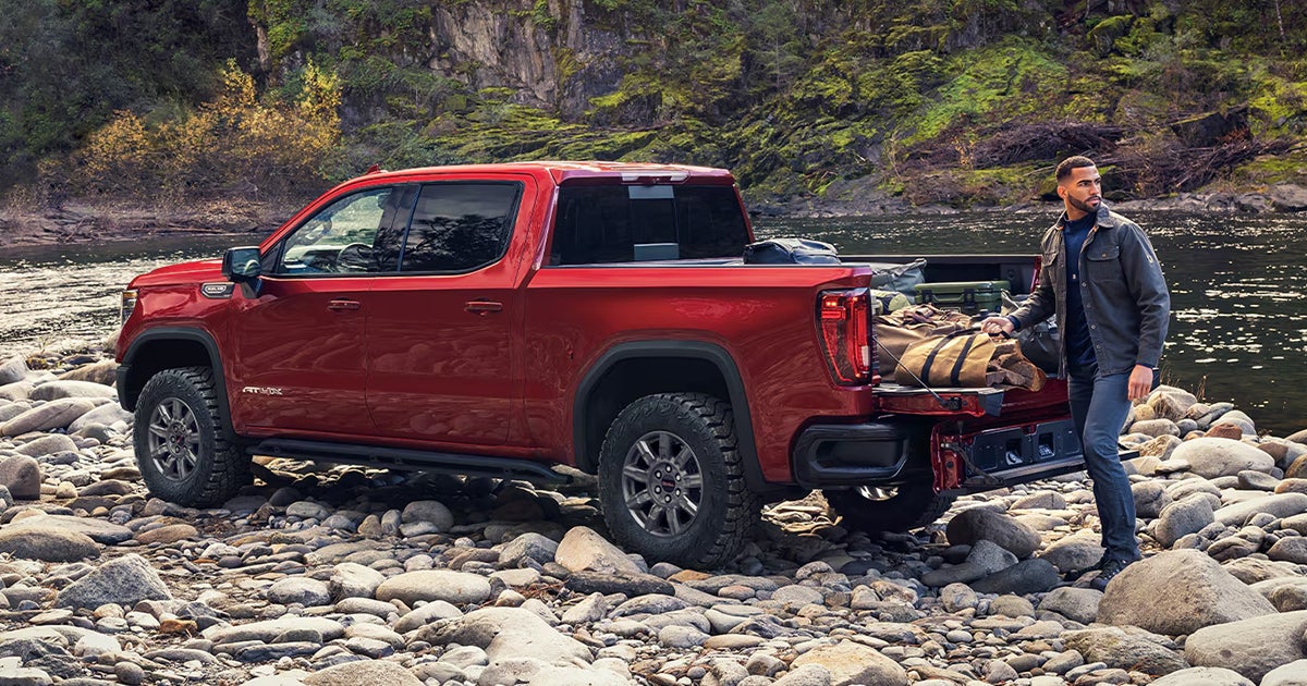 A red 2026 GMC Sierra 1500 pickup truck parked off-road by a river with the owner packing camping gear in the truck's bed.
