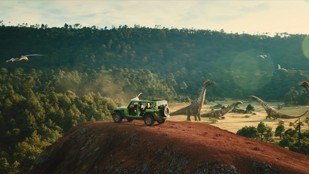 Jeep on top of a red rock with dinosaur in the background - image