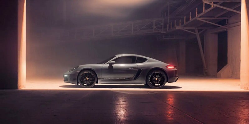 A silver 2024 Porsche 718-Caymn parked in a desolate warehouse with one overhead light illuminating the vehicle