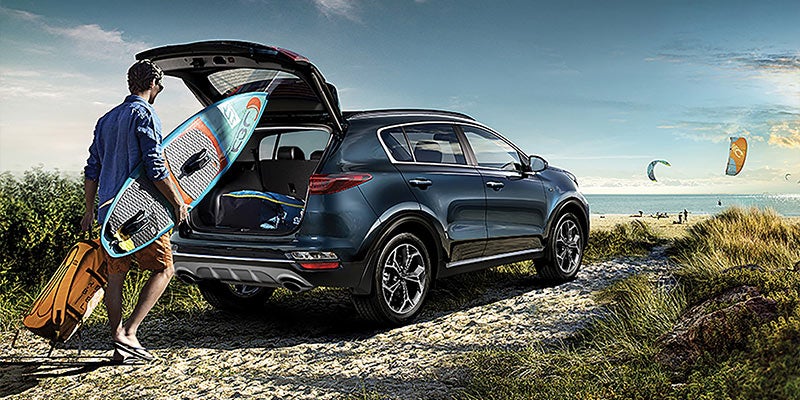 A man loading his paddle board and golf bag into the trunk of a blue 2022 Kia Sportage that is parked on a Hollywood, Florida beach