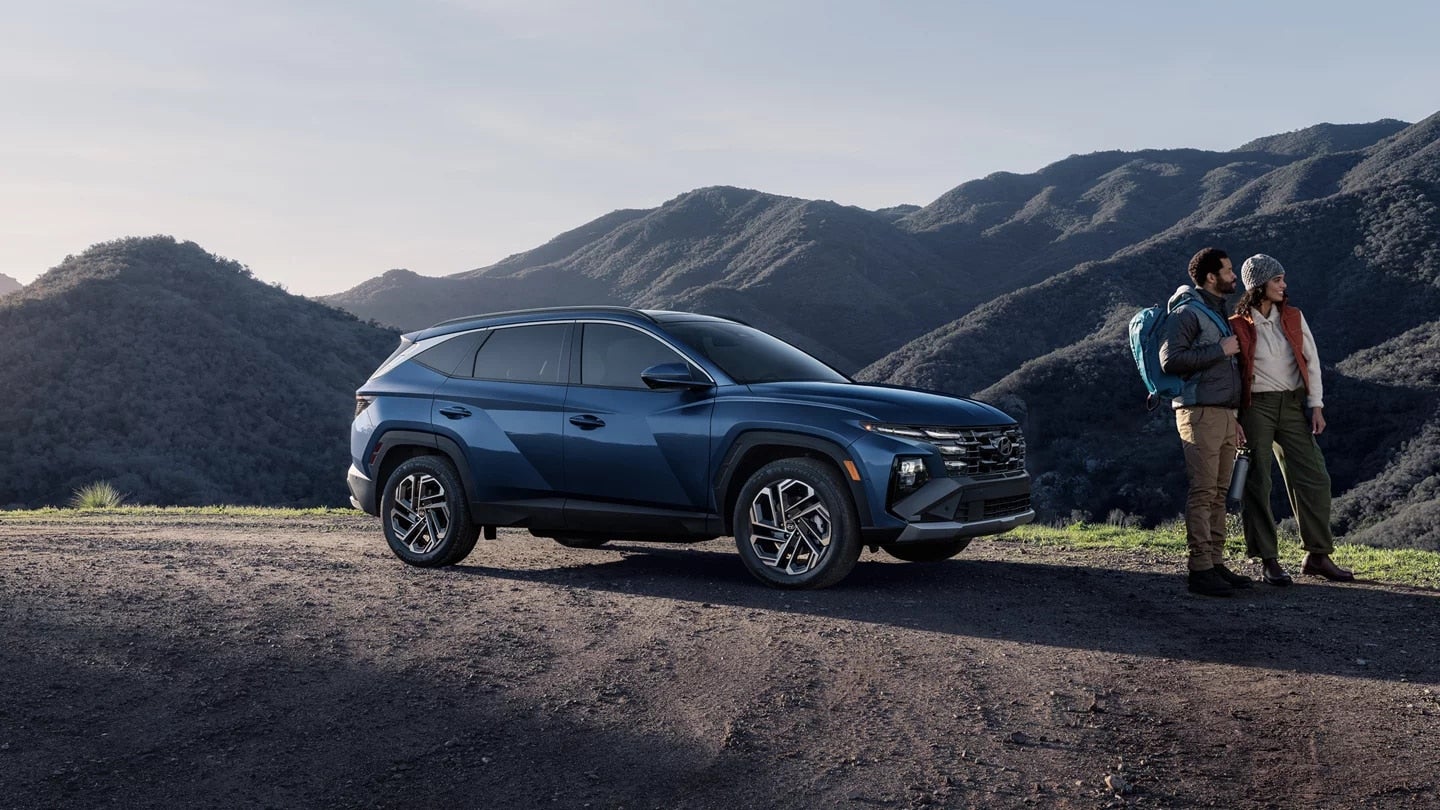 A blue-green SUV sits on a dirt road, facing slightly left, with mountains behind it under a bright sky.