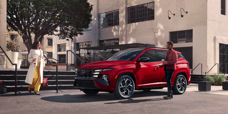 A red Hyundai Tucson parked on asphalt. A woman in a white coat approaches from the left, a man holds coffee on the right.