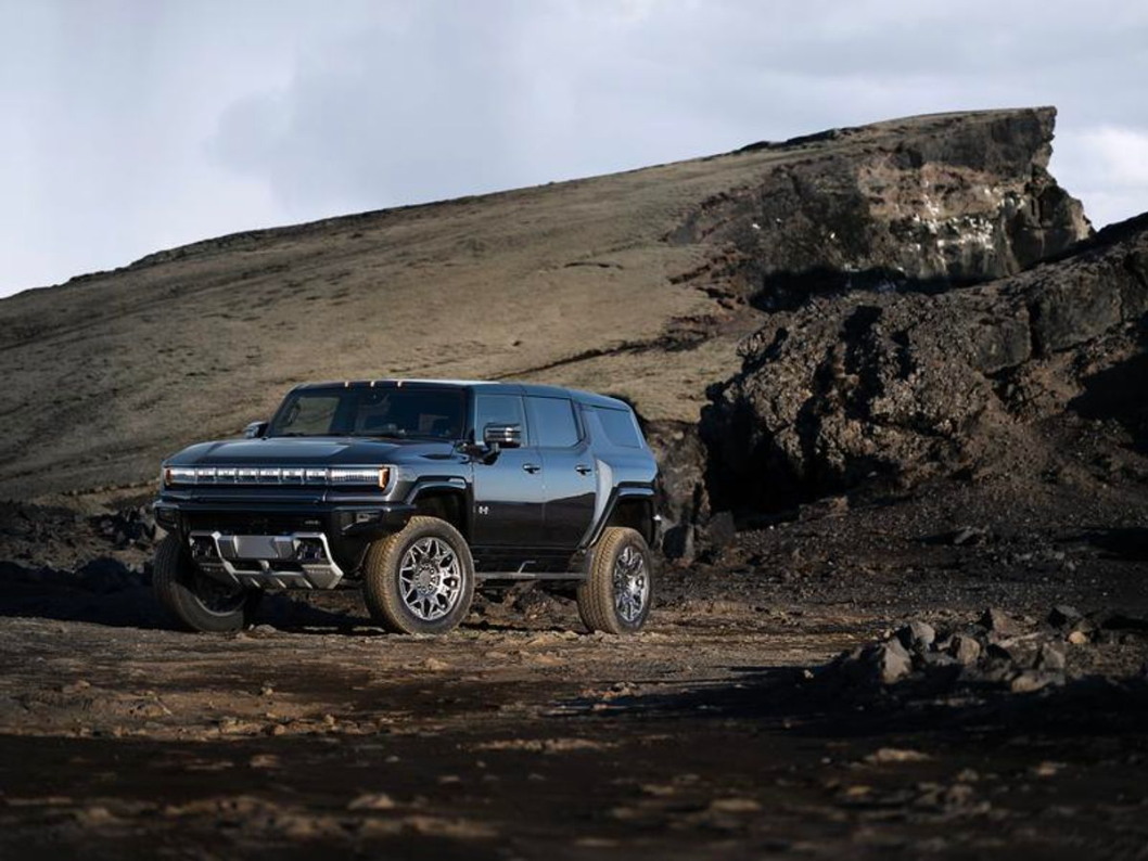Front driver's side view of a black 2025 Hummer EV SUV parked on rocky terrain with a mountain behind and blue skies overhead on a sunny day