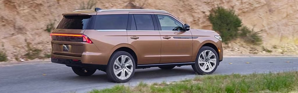 A bronze luxury SUV driving along a paved road next to a rocky hillside.