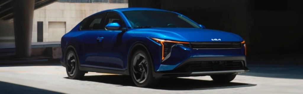 A blue 2025 Kia K4 sedan driving under a modern, urban structure.