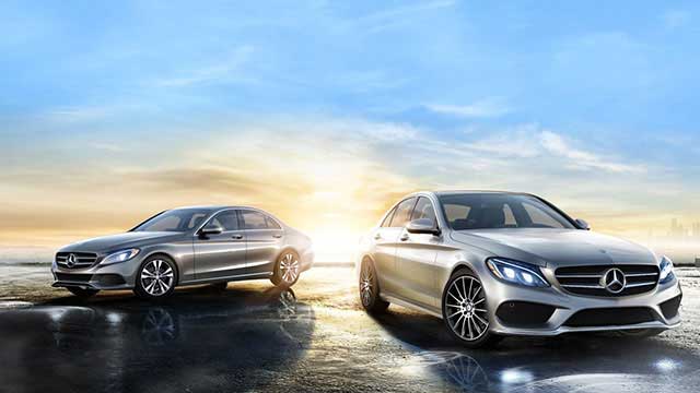 Two silver Mercedes-Benz luxury sedans parked on a reflective surface at sunset with a city skyline in the background.
