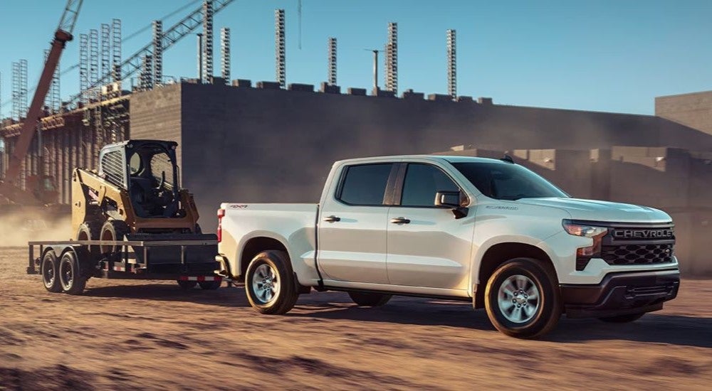 A white 2023 Chevy Silverado 1500 is shown towing construction equipment after leaving a Chevy dealer.