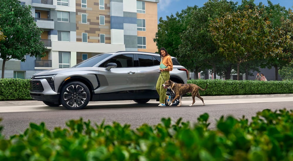 A woman walking a dog beside a parked white 2025 Chevy Blazer EV RS.