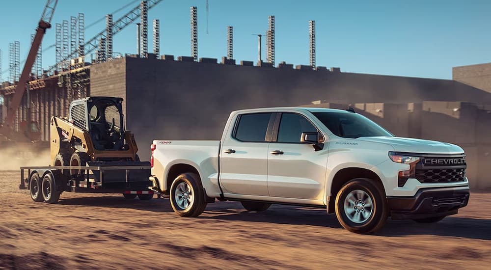 A white 2023 Chevy Silverado 1500 for sale is shown towing a skid-steer.