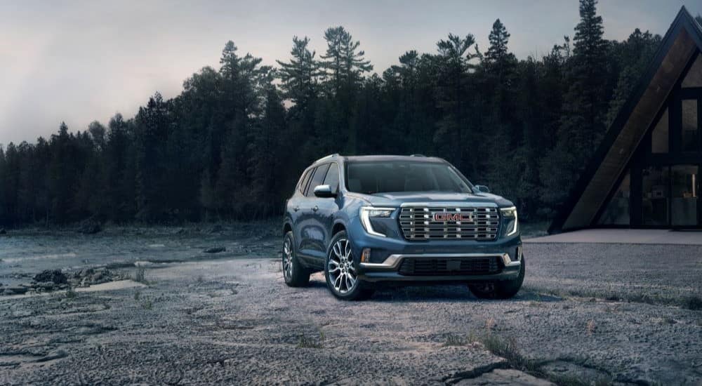 A blue 2024 GMC Acadia Denali is shown parked near a lake and cabin after leaving a GMC dealer.