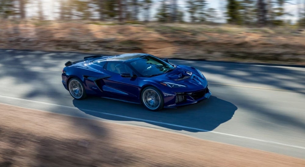 A dark blue 2025 Chevy Corvette ZR1 is shown driving on a tree-lined road.
