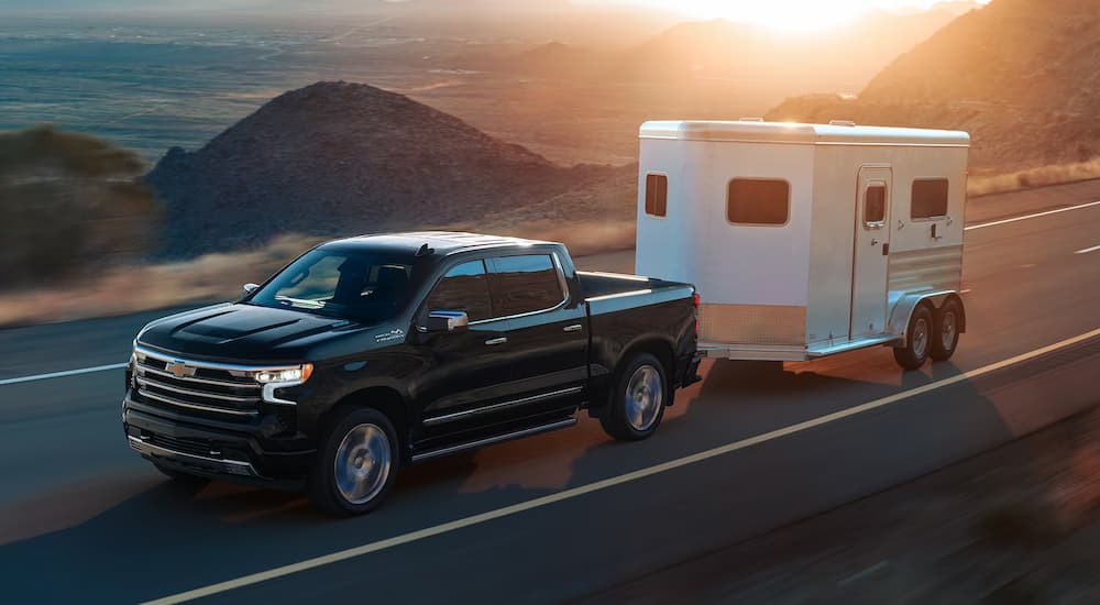 A black 2024 Chevy Silverado 1500 High Country towing a small trailer while driving to a Chevy dealer near Clinton Township.