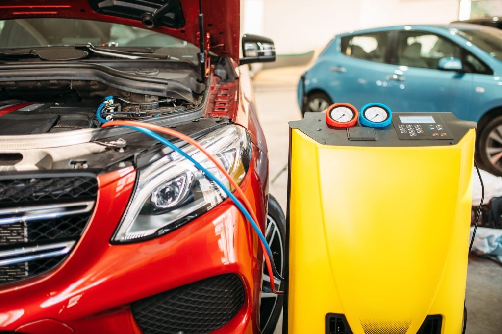 A red car with its hood open undergoing AC maintenance in a workshop, connected to a yellow air conditioning service machine.