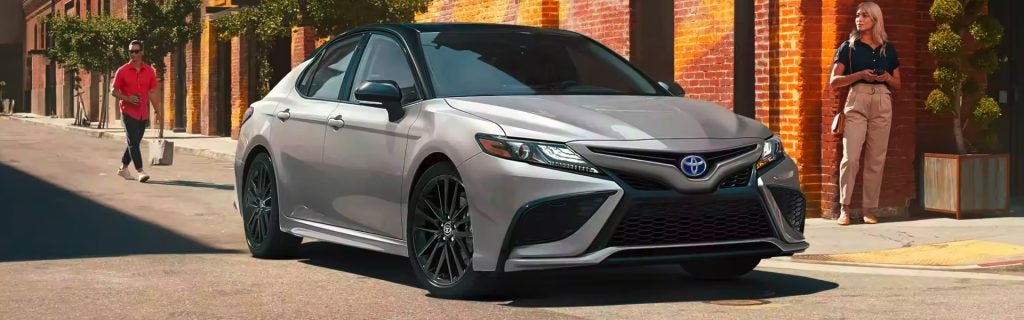 2025 Toyota Camry Hybrid driving through a city street with pedestrians and brick buildings in the background.