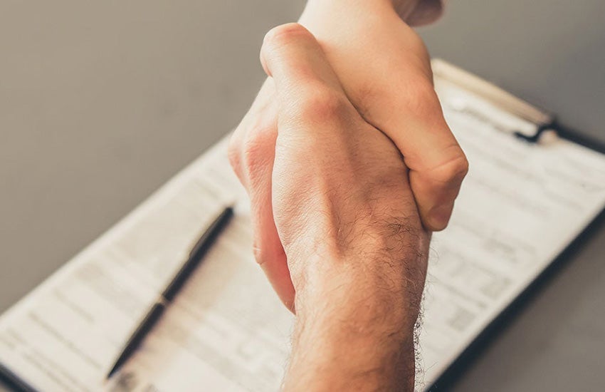 A close-up of a handshake from above, with a contract and clipboard on the table beneath, a pen laying atop the clipboard.