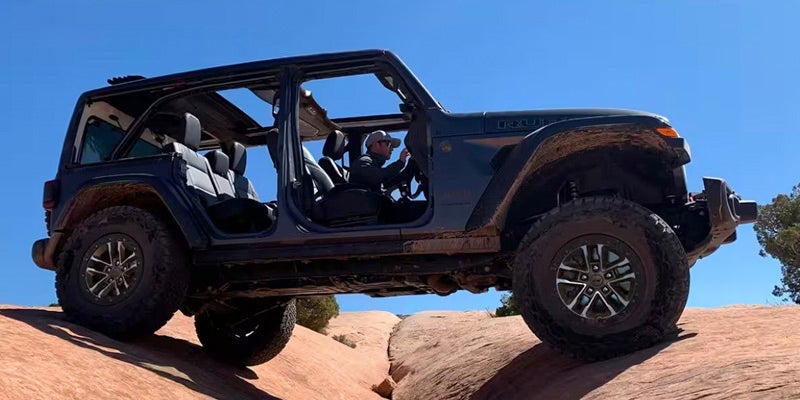 A blue Jeep Wrangler sitting atop a sand dune in the desert with its doors and roof removed, against a clear blue sky.