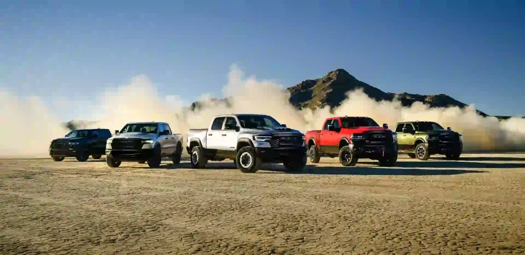 A lineup of five powerful Ram pickup trucks parked across a dry, cracked desert landscape, kicking up a massive cloud of dust behind them. The rugged trucks are displayed in various colors—black, white, silver, red, and olive green—with a rocky mountain peak visible under a clear blue sky in the background.