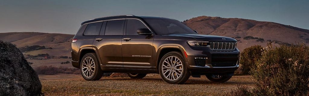 A sleek, dark metallic Jeep Grand Cherokee L parked on a gravel trail during golden hour, showcasing its three-row profile and multi-spoke wheels. Rolling hills and a warm sunset sky create a scenic backdrop behind the luxury SUV.