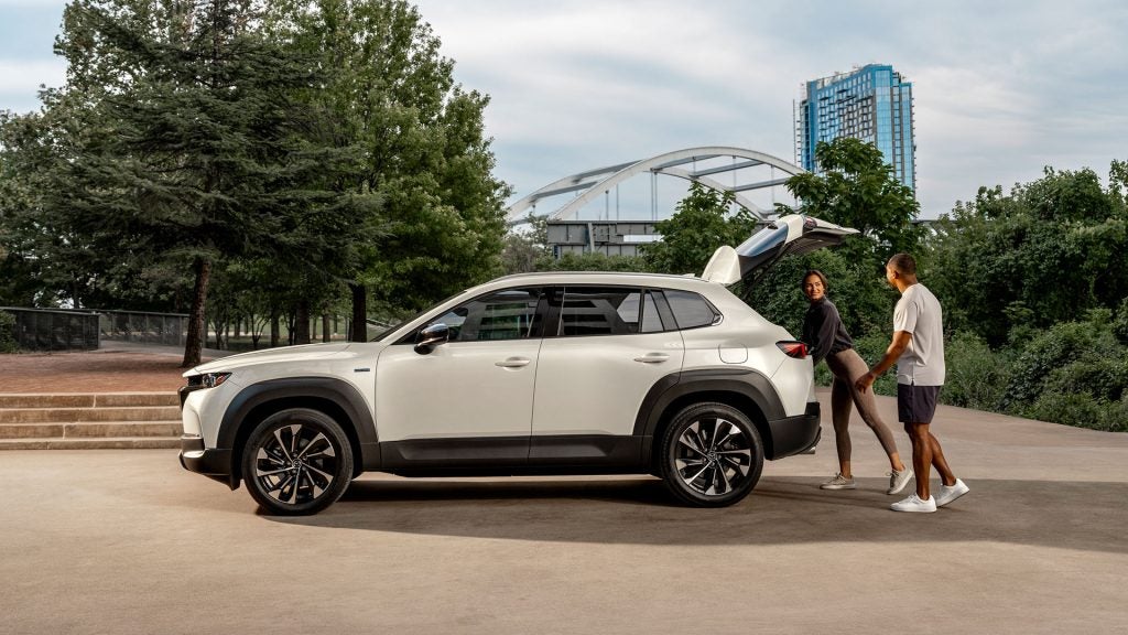 A family loading up the tailgate of a white Mazda CX-50 sitting in a city public space backed by trees and parkland.