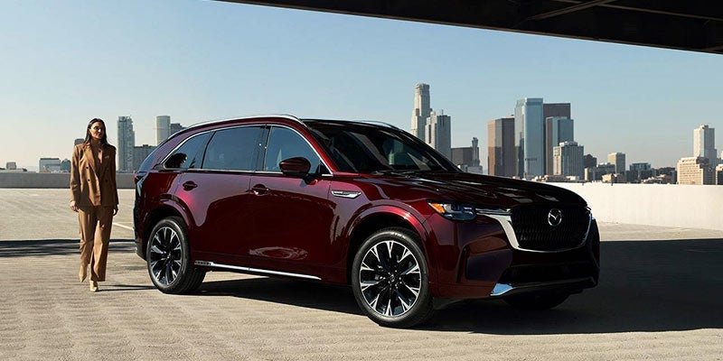 A dark metallic red CX-90 sitting on a tarmac with a woman standing near the rear of the vehicle. 