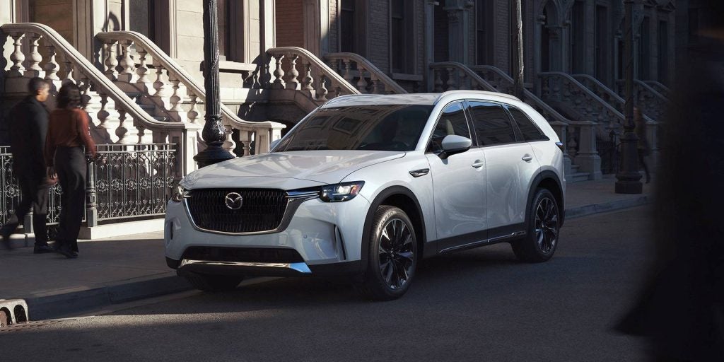 An image of a silver Mazda CX-90 sitting in front of a city apartment building.