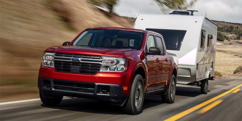 A red 2024 Ford Maverick towing a camper trailer. The road lines and background are blurred to give the impression of motion. 