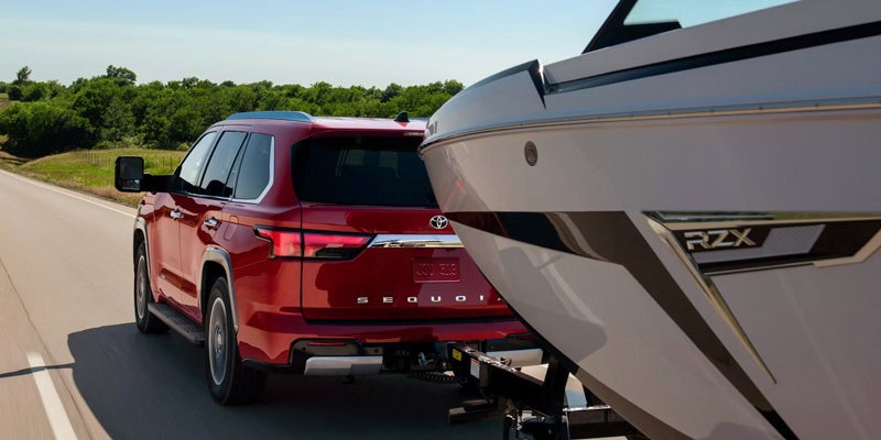 A red 2025 Toyota Sequoia towing a white and grey boat on a sunny road surrounded by greenery.