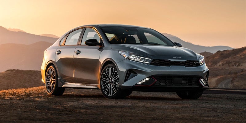 An image of a Kia Forte driving down the road with the setting sun and mountains behind. 