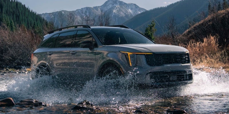 A silver Kia Sorento off-road, fording a stream in mountainous terrain.