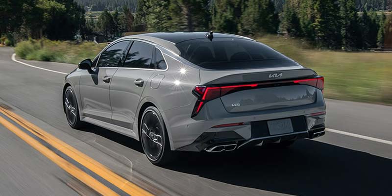 a 2026 gray Kia K5 sedan exterior shown from the rear and driver's side driving down a road in Shiloh, IL