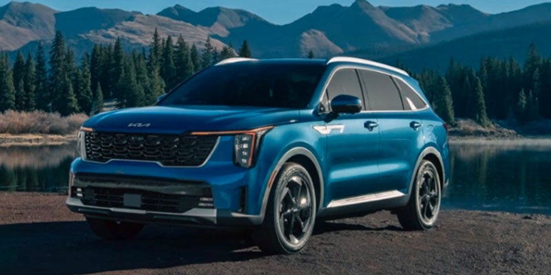 A metallic blue Kia Sorento Hybrid sitting in natural surroundings, backed by mountains, an evergreen forest, and a clear mountain lake. 