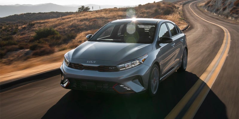 A gray Kia Forte showing the front and driver's side driving down a road