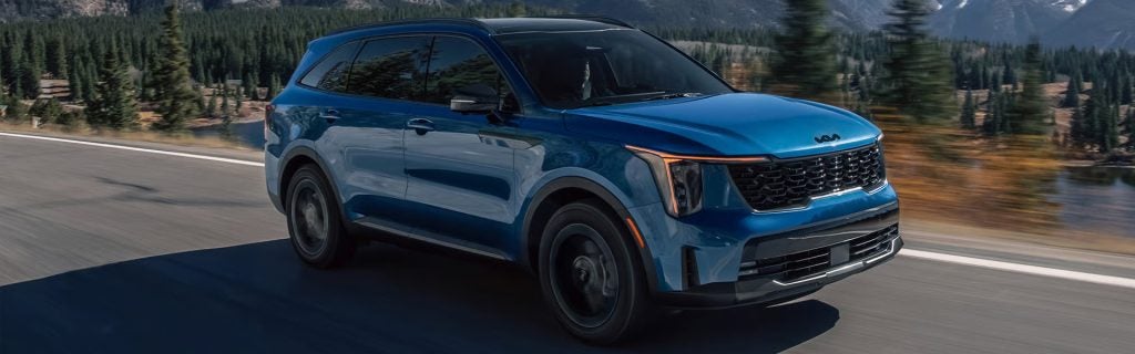 Blue Kia SUV driving along a mountain road with scenic forest and lake background.