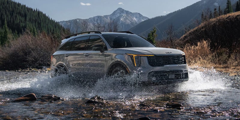 A silver Kia Telluride SUV splashing through a shallow river, with mountains and pine trees in the background.