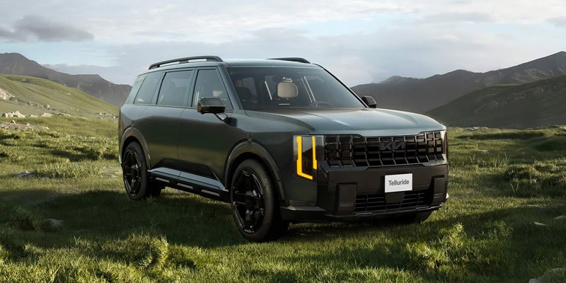 A charcoal gray Kia Telluride SUV parked on a grassy hillside with mountains under a cloudy sky.