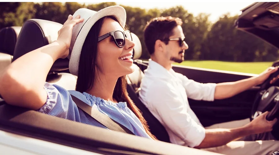 A smiling woman in a hat and sunglasses rides in a convertible with a man driving next to her.