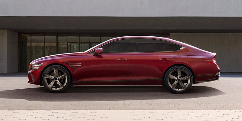 An all-new red 2025 Genesis G80 parked in front of a concrete industrialist building. - Genesis of Baton Rouge, LA