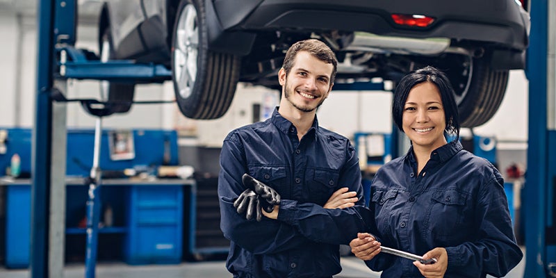 Two service technicians smiling and standing in a service center with a vehicle on a lifted bed ready for service behind them.