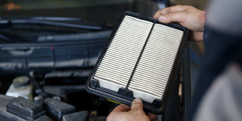 A close-up of a service technician holding a new clean air filter over a vehicle's engine.