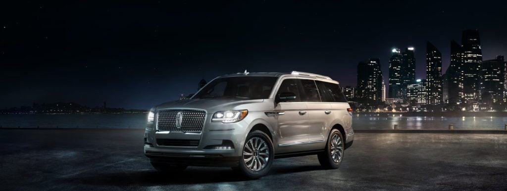 Interior view from the backseat of a 2024 Lincoln Navigator, showcasing the driver's seat. The vehicle is parked outside a modern home with warm lights glowing through the glass windows. A tree-lined landscape is visible through the driver's window. The interior features cool beige leather upholstery with elegant wood trim, and a large touchscreen display is prominently displayed.