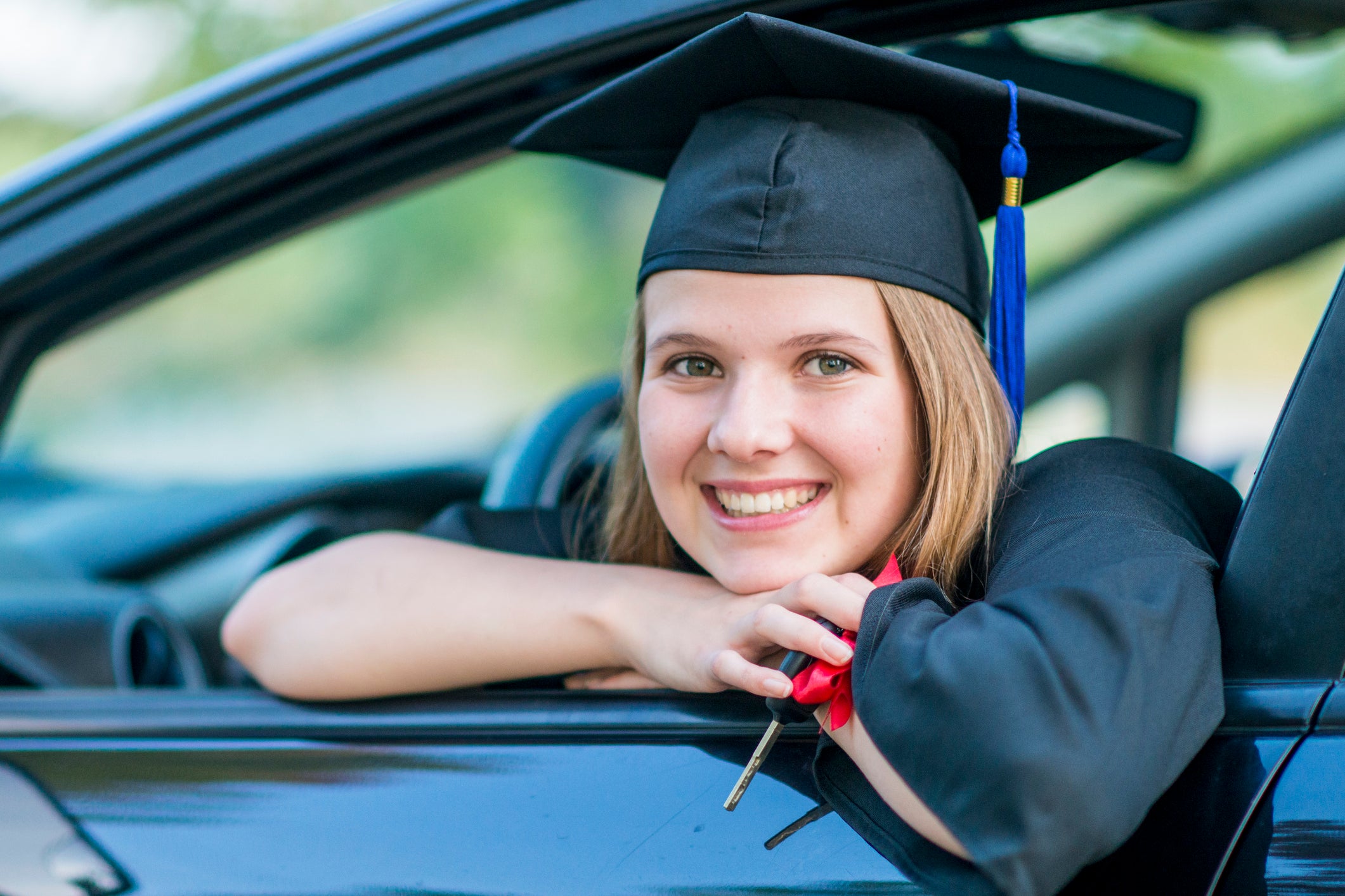 A Caucasian girl is sitting in a new car. She is wearing a graduation robe and cap. She is smiling out the window while holding her keys.