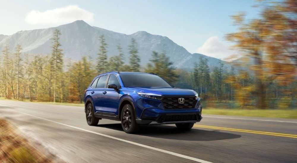 A blue 2024 Honda CR-V is shown driving by trees and mountains.